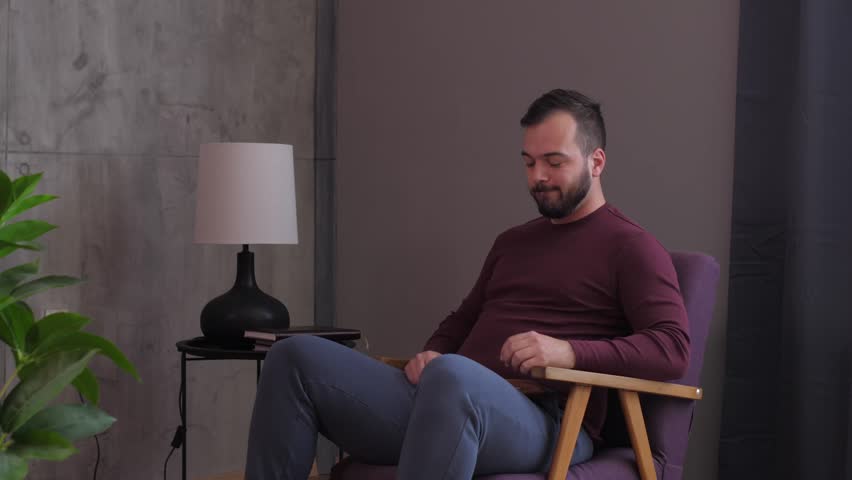Young man sitting in a cozy armchair puts on his eyeglasses and starts reading a book at home