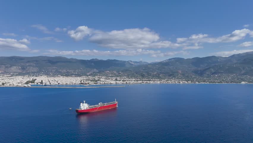 Aerial drone footage showing a red commercial cargo vessel anchored in the Messinian Gulf near Kalamata, Peloponnese, Greece. The ship rests on calm Mediterranean waters while the city of Kalamata stretches along the coastline beneath the rugged Taygetus mountain range. Harbor infrastructure, blue sea, scattered clouds and clear visibility create a scenic combination of maritime transportation, urban coastline and Greek landscape. Suitable for themes involving shipping, logistics, tourism, marine industry, Mediterranean destinations and international trade.