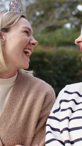 Vertical video: Laughing after facing pal in stripes, woman turning on park bench with Merry tiara. Outdoor, nature, friendship, laughter, portrait, candid, vertical video