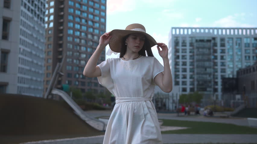 Woman in white dress holding a straw hat in a modern city plaza