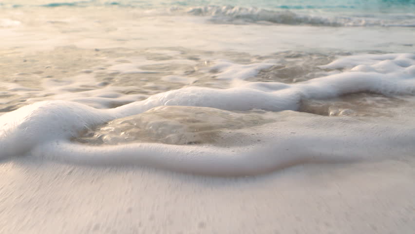 CLOSE UP: Shallow tidewater swirls around pale seashells that rest on sandy beach, leaving delicate foam and glossy reflections behind. Gentle motion of the sea on remote tropical shore in Indonesia.