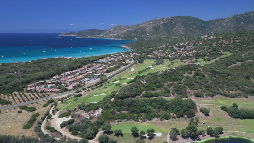 Drone footage of a coastal golf course overlooking a turquoise bay, rugged mountains, and seaside villas in Villasimius, Sardinia, Italy