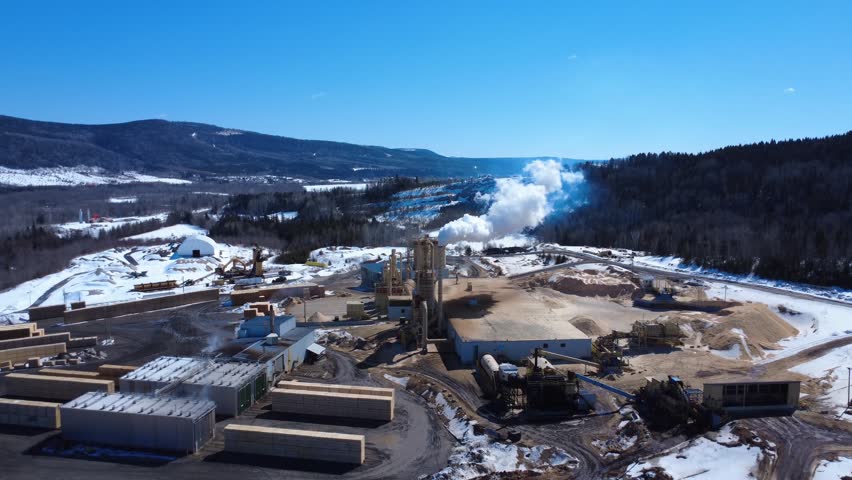 Industrial wood processing plant surrounded by snow, with prominent bag houses, lumber piles, and rural Canadian scenery.