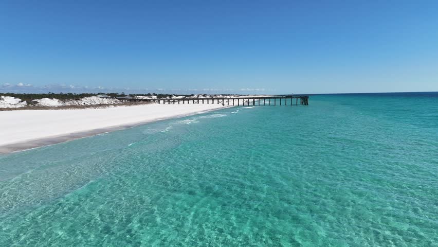Dynamic drone fly over turquoise Gulf waters gently lap against the white sandy beach near the wooden pier at St. Andrew State Park Pier, Panama City, Florida, US