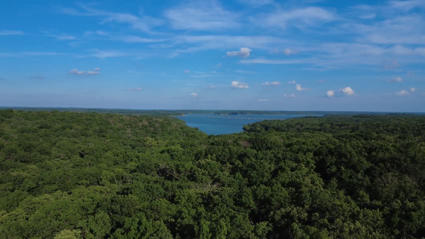 Drone footage above Ozark forest overlooking Stockton Lake, Missouri, with a long bridge, blue water, wooded shoreline, and summer sky.