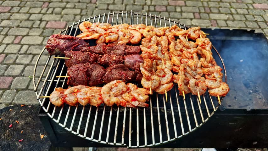 Close up of seasoned shrimp skewers and beef meat grilling on round barbecue grate outdoors