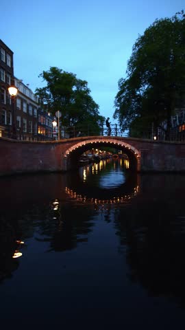 Immersive POV boat ride through Amsterdam canals under illuminated bridges at twilight	
