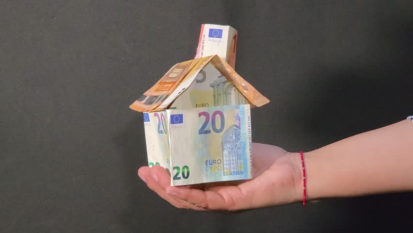 Close-up view of a woman hand holding a model house made of euro banknotes and turning it from side to side over a black background