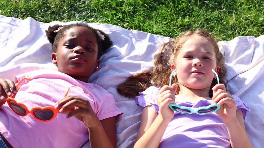 Top down view of two diverse little girls, one african american and one caucasian, lying on a blanket on the grass and putting on heart shaped sunglasses