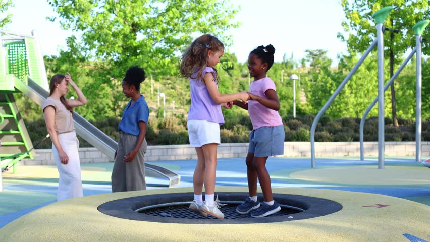 Two diverse young girls holding hands and jumping together on an in ground trampoline. Happy children playing in a public park on a sunny day