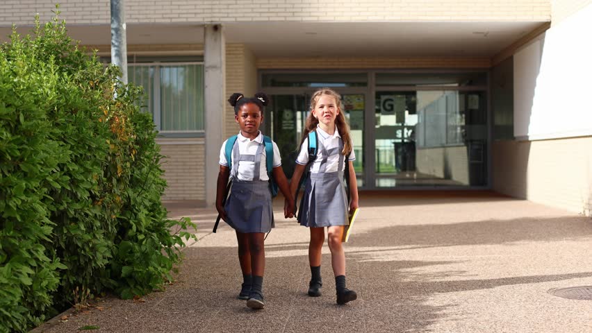Two smiling multiethnic girls in uniform with backpacks holding hands and walking together after classes. Elementary school student friendship