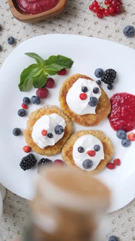 Delicious cottage cheese pancakes decorated with sour cream and fresh berries to resemble funny faces, getting sprinkled with cinnamon for a creative and healthy children