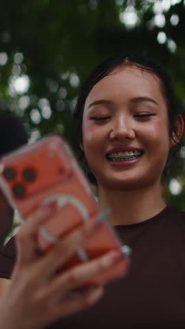 Happy young Asian woman with dental braces smiles while looking at her smartphone in a park