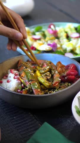Close up of a person eating a nutritious and colorful roasted vegetable salad bowl using chopsticks, enjoying a delicious and healthy vegetarian meal at a restaurant table for lunch or dinner