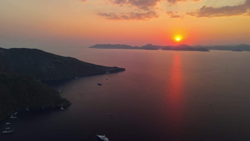 Aerial view of a Mediterranean bay at sunset, featuring distant islands, calm sea, anchored boats and golden reflections across the water beneath a colorful evening sky and mountain silhouettes.