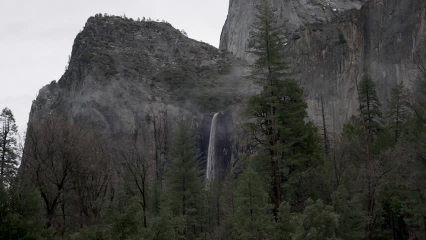 Scenic view of a waterfall flowing down a rocky mountain side surrounded by pine forest and fog in Yosemite