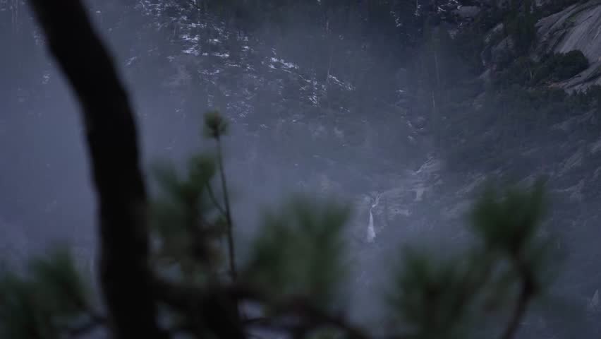 Scenic view of a waterfall flowing down a rocky mountain side surrounded by pine forest and fog in Yosemite