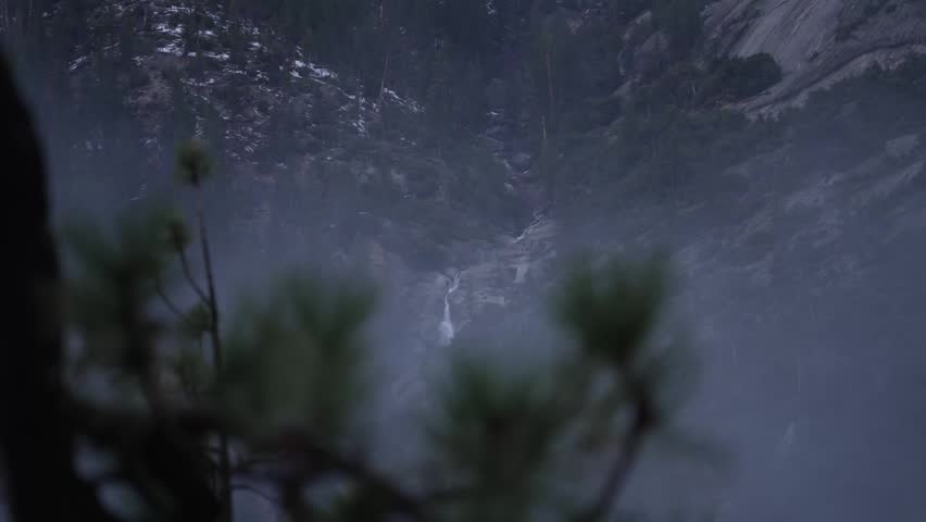 Scenic view of a waterfall flowing down a rocky mountain side surrounded by pine forest and fog in Yosemite
