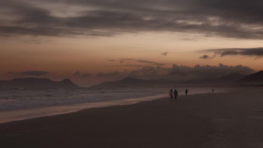 People stroll near the surf break in the late afternoon as the sun sets in golden light.