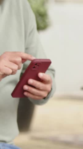 Vertical video: Tapping man in green top checking texts at home, camera panning showing red phone. Smartphone, interaction, scrolling, lifestyle, livingroom, window, vertical video