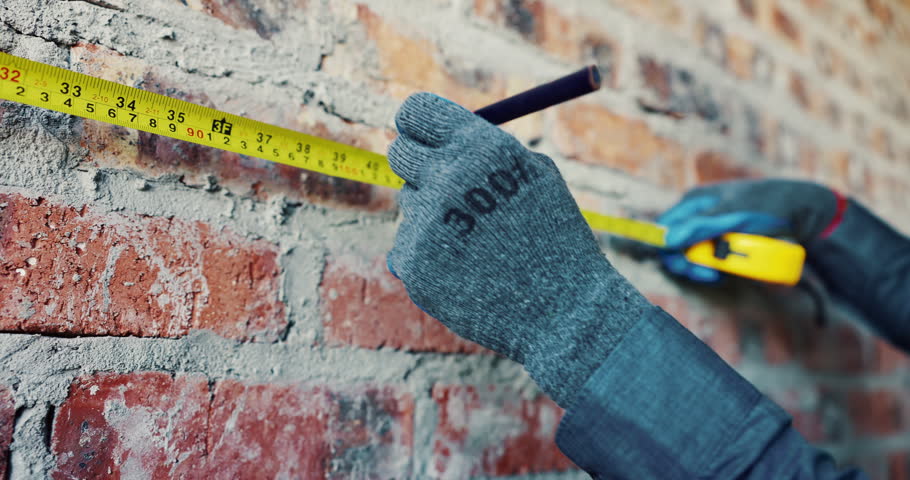 Hands, wall and tape measure at construction site for precision, dimension and layout for building. Man, gloves and tools or writing for measurement, structure and property development or renovation