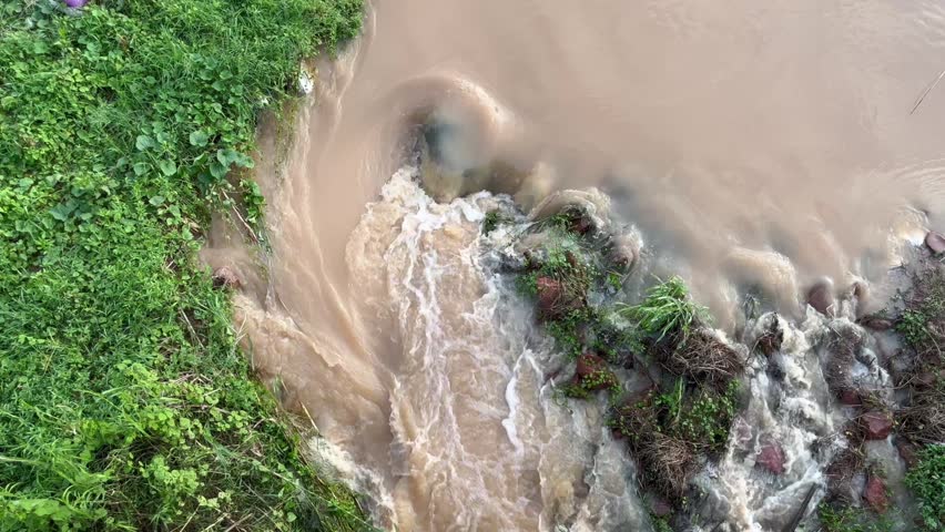 powerful muddy flash flood current hitting riverbank and rocks, demonstrating severe water erosion, natural disaster, and environmental crisis context