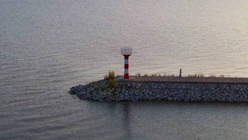 Aerial view of Sayram Lake in Xinjiang, China, featuring a red-and-white lighthouse on a rocky jetty during golden hour, with tourists walking along the shore.