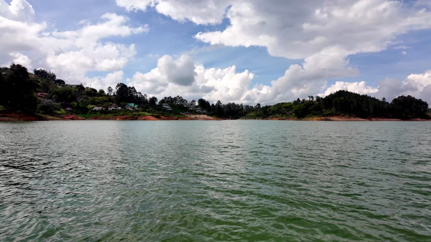 A boat perspective approaching the stone cross marker that signifies the submerged ruins of Old El Peñol town within the Guatape reservoir on a bright, sunny day.
