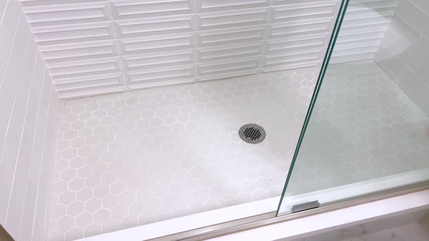 A close up of a clean white shower floor covered with hexagon tiles. The shot focuses on the tile texture and stainless steel drain under bright lighting.
