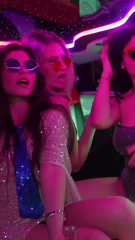 Glamorous multicultural women wearing sparkly dresses and heart shaped sunglasses having fun, drinking, and dancing together inside a limo with vibrant pink neon lights for a special occasion
