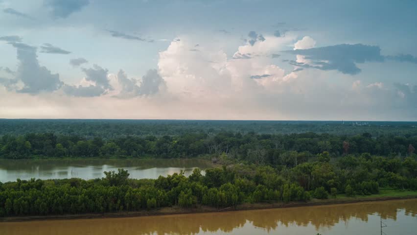 Aerial timelapse showing the muddy Yazoo River beside Vicksburg, MS, with dense forested wetlands, calm water reflections and a soft cloudy sky. Barges drift by on the Mississippi River trade route.