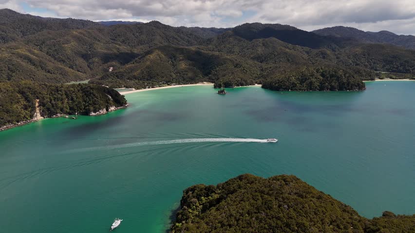 Drone view of small white boat leaving long wake in turquoise ocean water in Abel Tasman National Park, New Zealand. Aerial