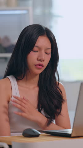 Girl with black hair in white shirt feeling shoulder pain while using laptop at desk in home workspace, tired woman showing neck discomfort, strain, injury during computer work Vertical