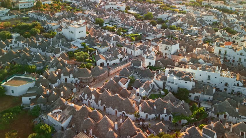 Aerial view of Alberobello during golden hour. The unique conical roofs and white stone dwellings are bathed in warm sunset light across the historic landscape of Puglia, Italy