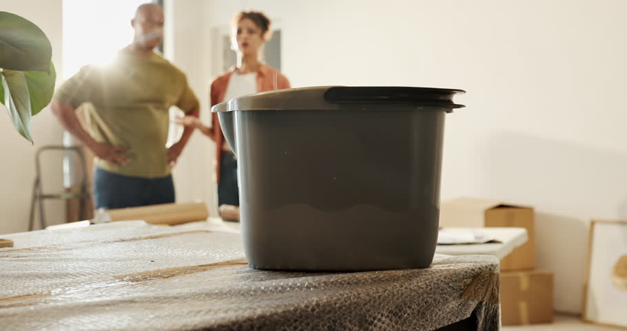 Leak, bucket and couple in new home with stress for repair, maintenance or assistance with moving. People, container and water dripping for plumbing emergency in living room with construction mistake