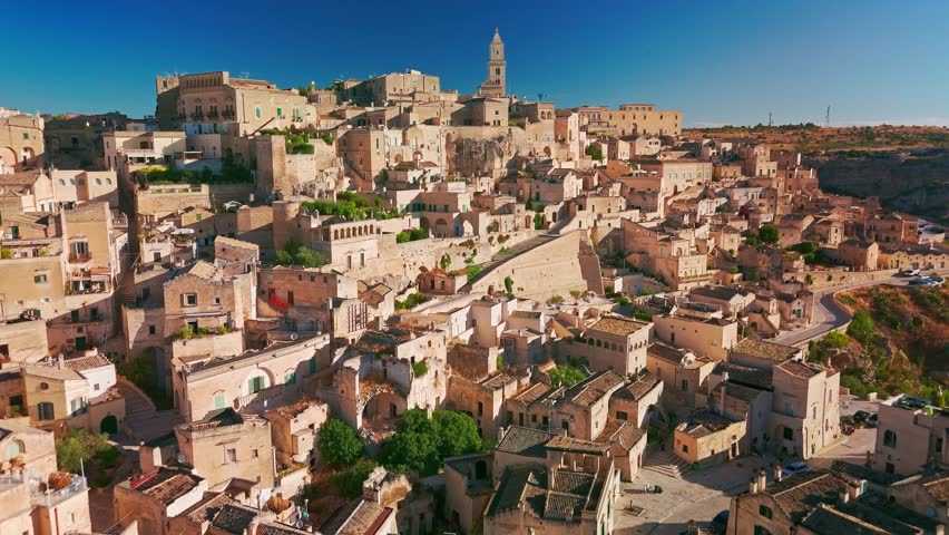 Drone shot of Matera under a bright summer sun. The clear blue sky highlights the white limestone Sassi dwellings and the Romanesque Cathedral in the historic landscape of Basilicata