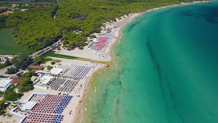 High-angle drone shot of a long white sand beach in Salento during August. Lots of colorful sun umbrellas and people fill the coast by the turquoise Ionian Sea in Puglia Italy.