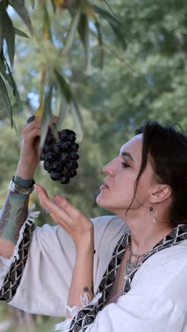 A beautiful brunette in a bohemian style relaxes outdoors in summer against a backdrop of a green forest, holding a large bunch of juicy black grapes