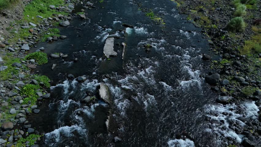 Aerial view of a fast river with rocky banks and green moss. White water rapids churn vigorously. Bird glides above the water. Close-up shows splashing waves. Wide shot reveals valley path. Ideal relaxing background