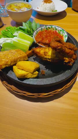 A portrait view of a plate of the Indonesian fried chicken, fried tofu, fried tempe, chili sauce (sambal), and vegetables with cucumber, chayore, cabbage, and kemangi leaves.