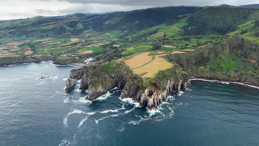 Rocky coastline of Azores with steep Atlantic cliffs, green farmland above and ocean waves breaking along shore