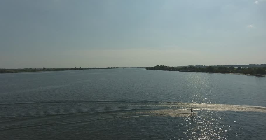 Aerial drone view of a motorboat towing a water skier across a wide sunlit river on a clear summer day. The boat trails a long foamy wake and a fan of ripples over the sparkling water, with low green banks, reeds, trees and riverside cottages along the shoreline. A dynamic tracking shot of summer water sports, recreation and active outdoor lifestyle, well suited to travel, tourism, leisure and adventure content.
