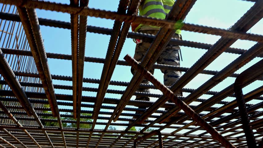 Low angle view of a construction worker walking across steel rebar reinforcement on a bridge or infrastructure site, showing civil engineering, safety equipment and structural concrete preparation.