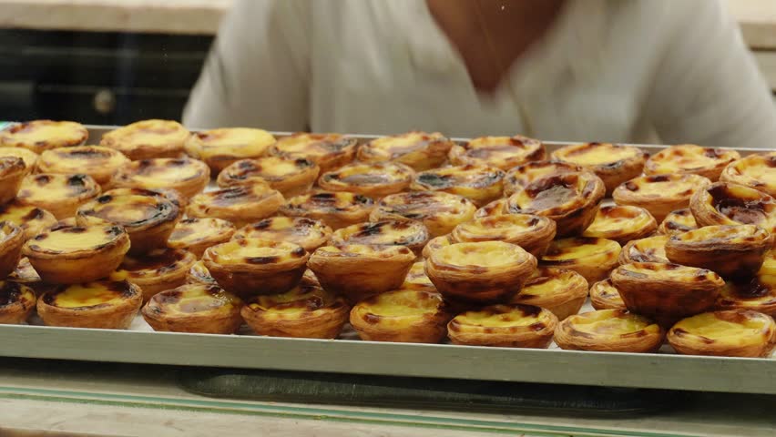 Large metal tray filled with traditional Portuguese custard tarts, pastel de nata, fresh out of the oven with caramelized tops on display at a local bakery counter.