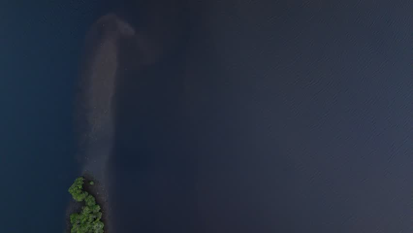 Aerial view captures a dark blue lake. Rounded island with dense green forest is centred. Water reflects the sky