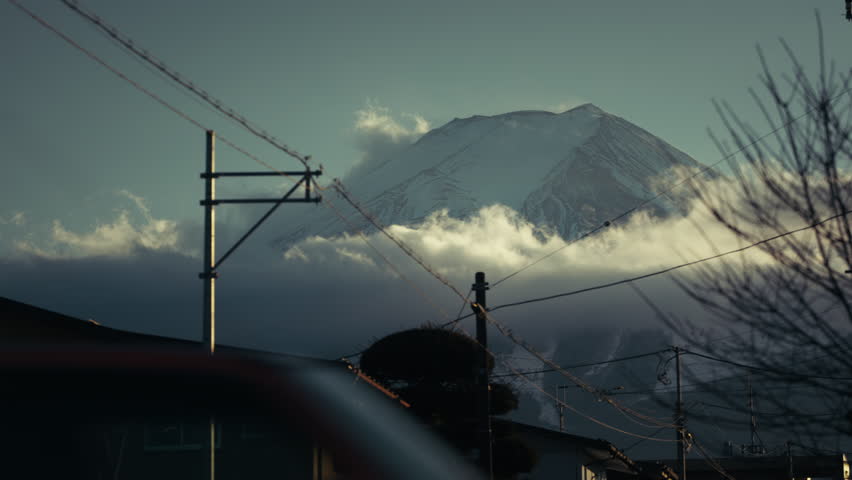 Cars passing through a Japanese town with snow-covered Mount Fuji emerging through drifting winter clouds in the background