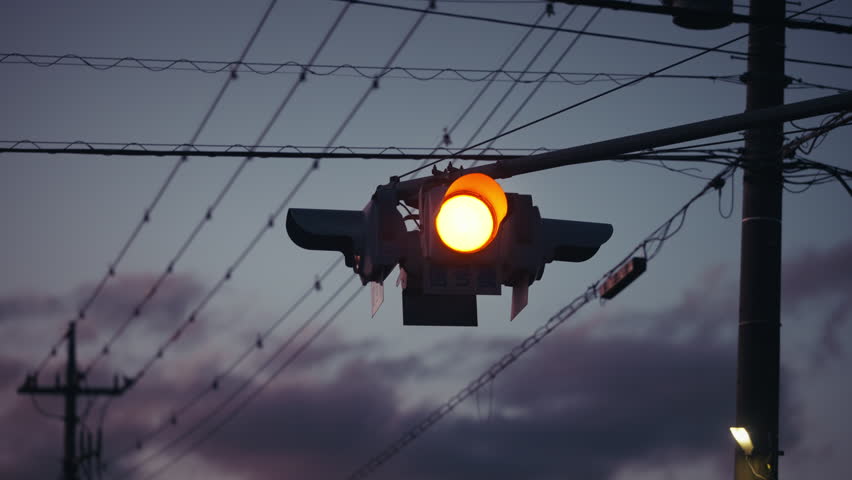 Blinking amber traffic signal suspended above a Japanese town at dusk with overhead power lines silhouetted against a dramatic cloudy evening sky