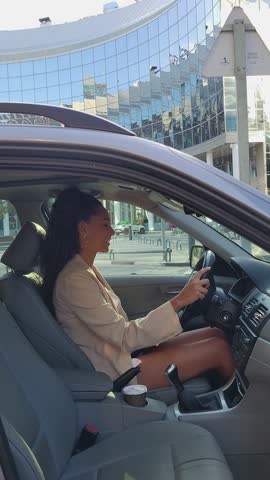 Confident young businesswoman driving her car in front of a modern glass building on a sunny day