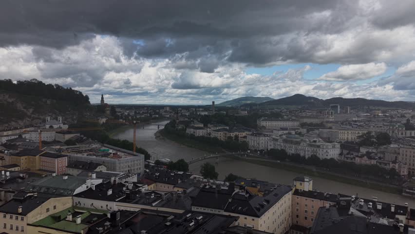 Drone footage moving toward the city center of Salzburg during a passing rainstorm. The Salzach River, historic buildings, and surrounding hills are framed beneath dramatic cloud formations and shifting light.