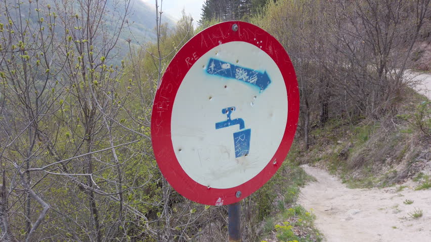 A sign indicates where to find a water source along a hiking trail in a forest. It shows an arrow pointing towards the left. The area has trees and a dirt path.